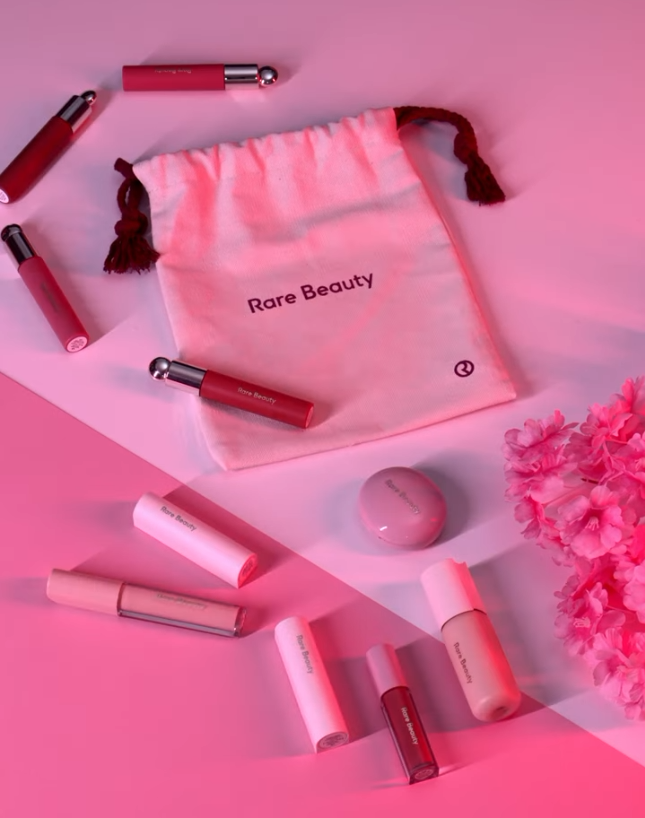 Rare Beauty cosmetics arranged around a branded pouch on a pink set.